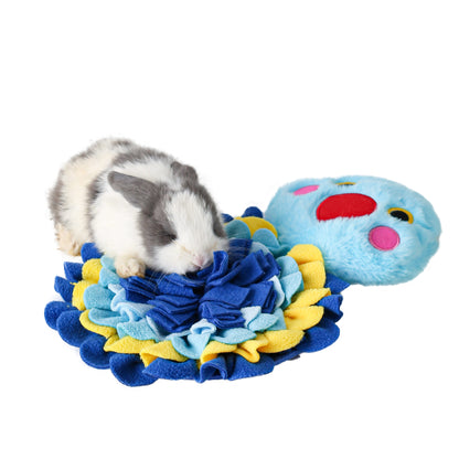 A small gray and white rabbit is sniffing a colorful plush rabbit toy with a blue, yellow, and red patterned fabric mat underneath, photographed on a clean white background.